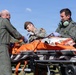 Three military personnel in green flight suits and combat uniforms are attending to a patient on an orange stretcher outdoors. The scene appears to depict a medical evacuation or emergency response exercise, with personnel wearing gloves and aviation headsets. The setting is a grassy outdoor area under clear blue skies.