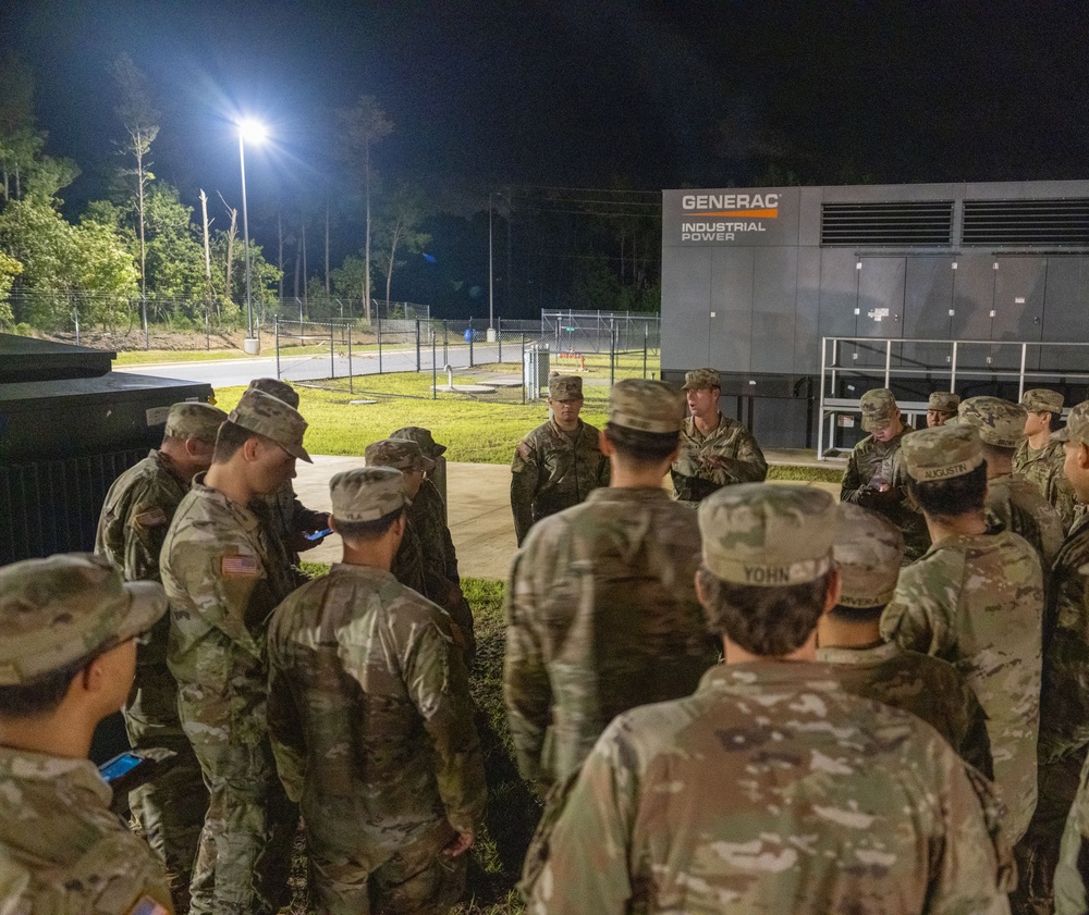 A group of military personnel in camouflage uniforms are gathered outdoors at night near a large Generac Industrial Power generator unit. They appear to be receiving a briefing or instruction from one or two individuals facing the group. The scene is illuminated by overhead floodlighting.