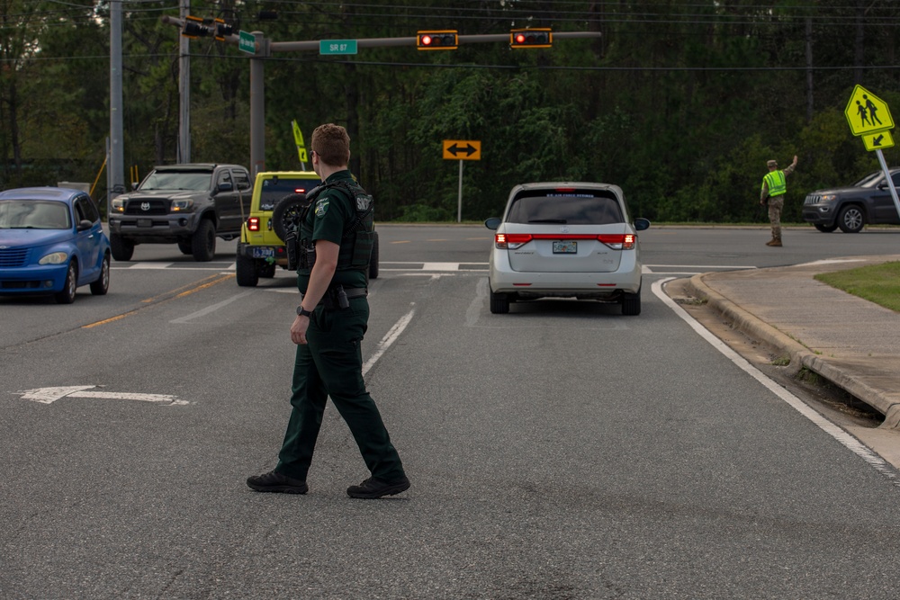 A law enforcement officer wearing a dark green sheriff's uniform and tactical vest is directing traffic at an intersection marked SR 87. A second person in a high-visibility vest is also visible in the background assisting with traffic control. The scene appears to be a managed traffic situation, possibly related to an emergency, evacuation, or school zone activity.