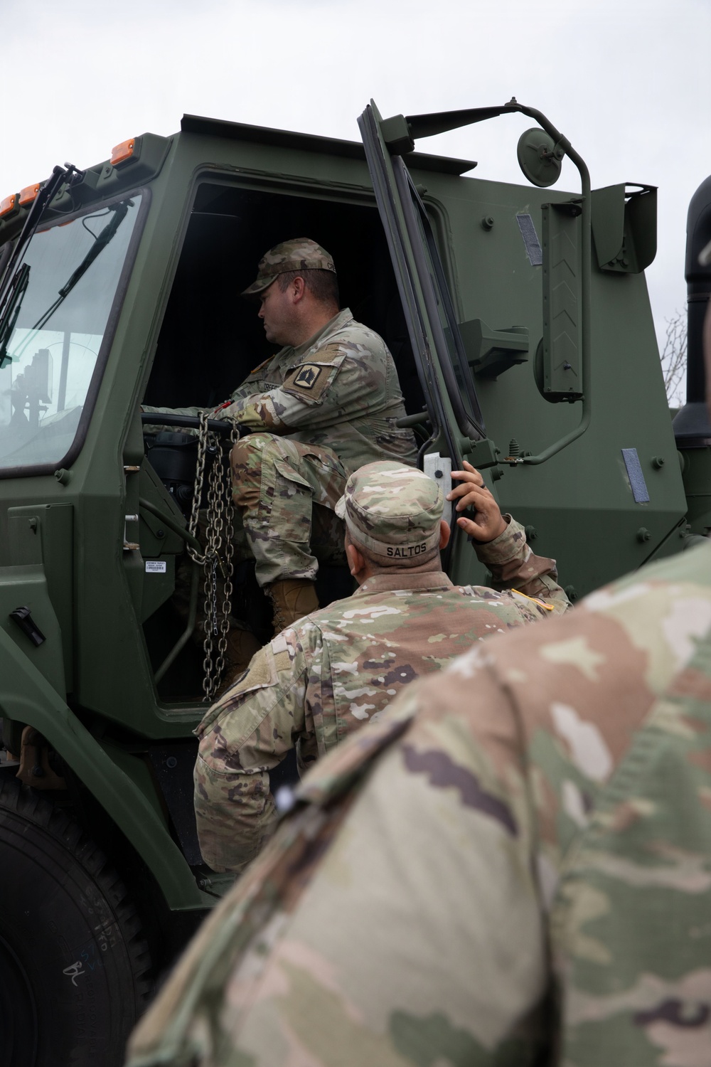 The image shows U.S. Army soldiers in Operational Camouflage Pattern (OCP) uniforms interacting with a large military tactical vehicle, likely an FMTV or similar heavy transport truck painted in olive drab green. One soldier is seated inside the open cab of the vehicle while another soldier, identified by the name tape 'SALTOS,' stands below holding what appears to be a device or document. A third soldier is partially visible in the foreground.