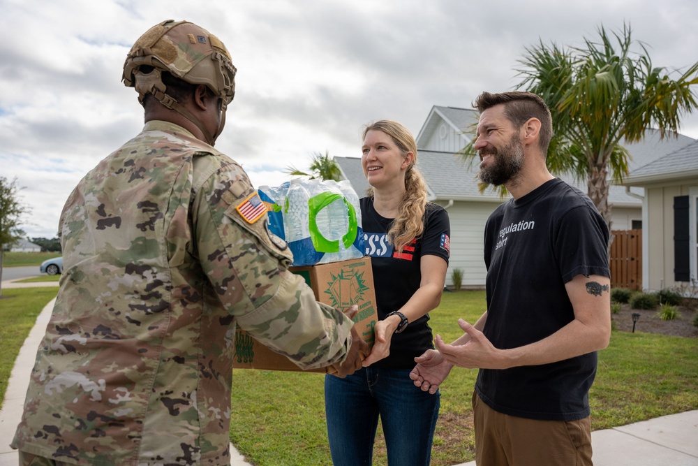 A U.S. Army soldier in OCP (Operational Camouflage Pattern) uniform is receiving or exchanging supplies — including a case of bottled water and a cardboard box — from two civilians outside a residential building. The interaction appears friendly and cooperative, suggesting a relief or community assistance operation. The setting is a suburban neighborhood with palm trees and overcast skies, consistent with a post-disaster or humanitarian aid scenario.