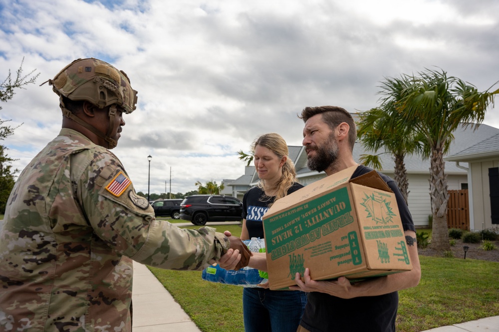 A U.S. Army soldier in full combat uniform and helmet is distributing emergency supplies to two civilians in a residential neighborhood. The man is receiving a cardboard box labeled 'Quantity: 12 Meals' (MREs) while the woman holds a pack of water bottles. The setting and supplies suggest a disaster relief or emergency response operation.
