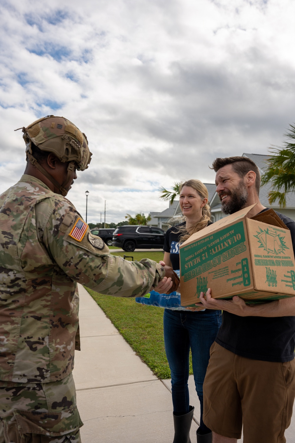 A U.S. Army soldier in full combat uniform and helmet shakes hands with a civilian man who is holding a cardboard box labeled 'Quantity: 12 Meals,' while a woman stands nearby smiling. The interaction appears to be a relief supply distribution or donation handoff, likely in the aftermath of a natural disaster. The setting is a residential neighborhood with palm trees and overcast skies suggesting a coastal or subtropical location.