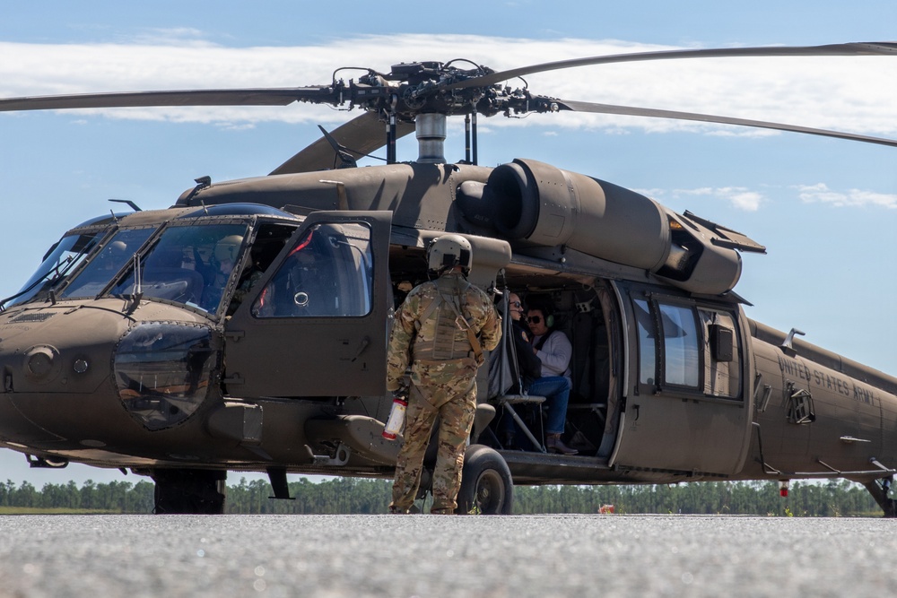 A United States Army UH-60 Black Hawk helicopter is parked on a tarmac or airfield. A soldier in full combat uniform and flight helmet stands at the open cabin door, interacting with civilian passengers seated inside. The scene appears to be a demonstration, orientation flight, or VIP transport event.