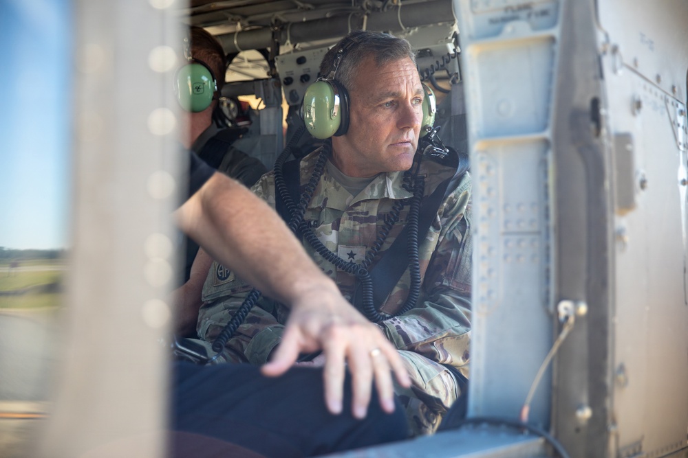 A military officer in camouflage uniform is seated inside what appears to be a military helicopter, wearing green aviation headphones with a communication cord. He is looking outward through the aircraft window with a focused expression. At least one other person wearing similar headphones is visible in the background.