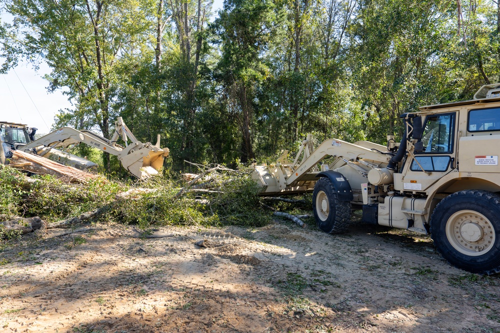 Two tan/beige military-style heavy equipment vehicles are engaged in debris clearing operations in a wooded area. The machines appear to be using front-loader attachments to move and pile fallen trees and brush. The scene suggests post-storm or disaster relief debris removal work.