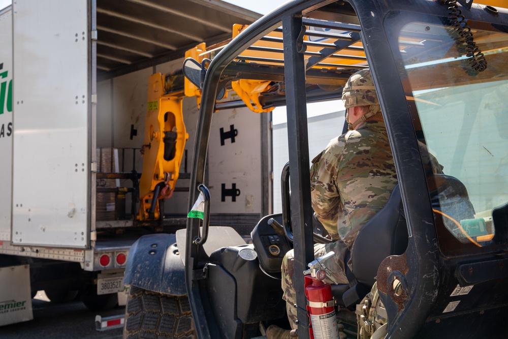 A military service member in MultiCam/OCP camouflage uniform and helmet is operating a forklift near the open rear of a commercial box truck. The forklift, which has a yellow attachment mechanism and a mounted fire extinguisher, appears to be engaged in loading or unloading operations. The setting appears to be an outdoor logistics or supply area.