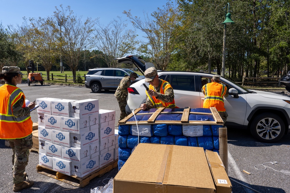Military personnel and workers in high-visibility orange safety vests are distributing emergency supplies at what appears to be a disaster relief distribution point in a parking lot. Boxes labeled 'SENT Emergency Drinking Water' are stacked on pallets alongside blue-bagged items on a wooden pallet. Vehicles are lined up to receive supplies, suggesting a drive-through relief operation.