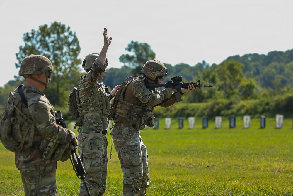 Three military personnel in multicam/OCP camouflage uniforms are on an outdoor firing range. One soldier is actively aiming a rifle (appears to be an M4 carbine) downrange at a row of targets, while a second individual stands beside them with one hand raised, likely acting as a range safety officer or instructor. A third soldier stands to the left holding a weapon in a relaxed position.