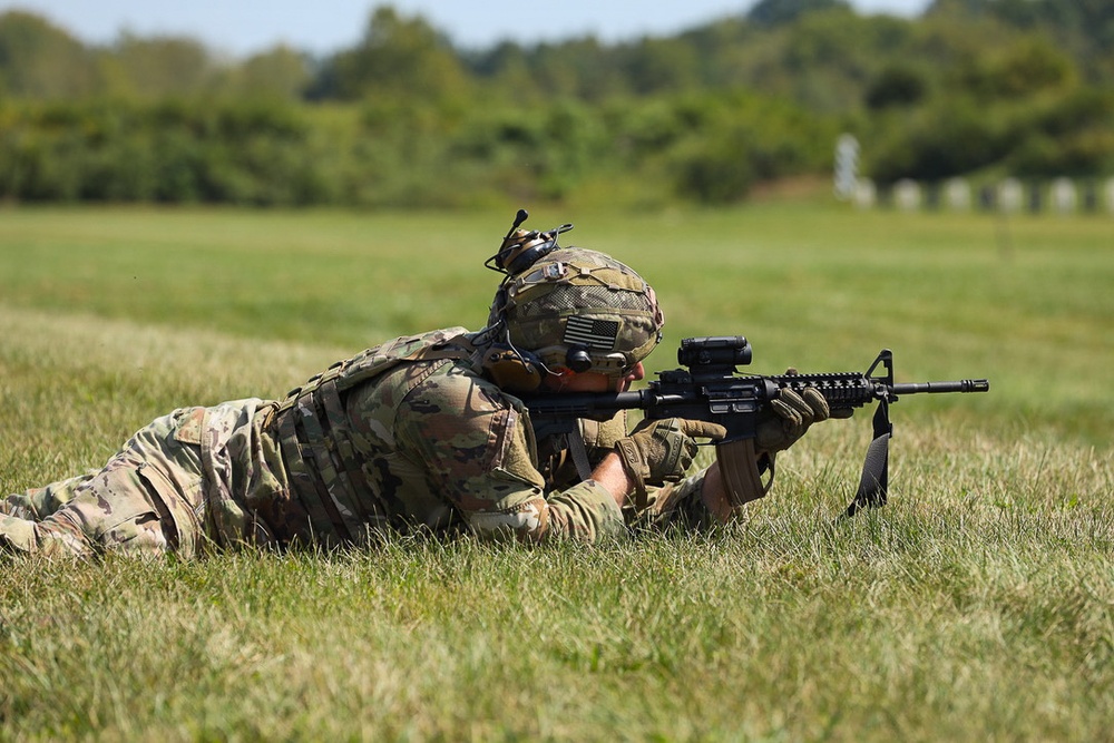A soldier in full combat gear is in a prone firing position on a grass field, aiming a rifle downrange. The soldier is wearing MultiCam/OCP camouflage uniform, a tactical helmet with mounted accessories, hearing protection, and a plate carrier vest. The background shows a open range field with trees and what appear to be target stands in the distance.