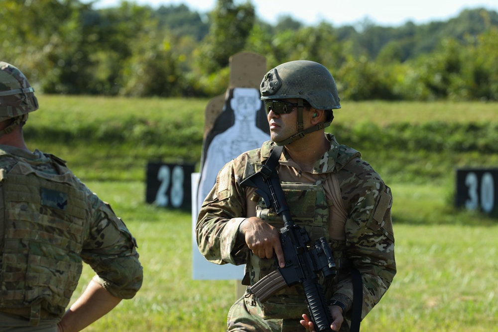 Two soldiers in full combat gear are at an outdoor shooting range during what appears to be a training exercise. The primary subject is holding an assault rifle in a relaxed carry position while standing near numbered target stands. The setting is a grassy range with silhouette targets visible in the background.