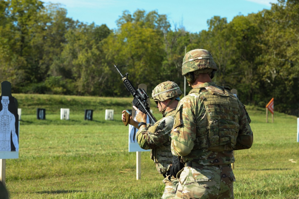 Two military personnel in full combat gear are at an outdoor shooting range. One soldier is holding and inspecting a rifle (appears to be an M4/M16-style carbine) while the other observes from behind. Multiple silhouette targets are positioned downrange on a grassy field with a tree line in the background.