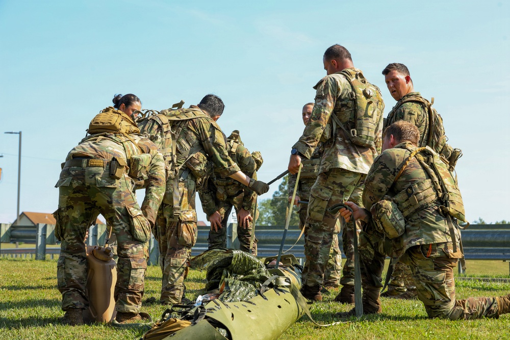 A group of military personnel in camouflage uniforms are gathered on a grass field, working together around what appears to be a litter or stretcher on the ground. The soldiers are wearing full combat gear including tactical vests and backpacks, and appear to be conducting a casualty evacuation or medical training exercise. The setting appears to be a military installation or training area.