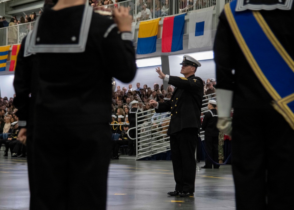 A military ceremony is taking place in a large indoor venue, with a naval officer in dress uniform conducting what appears to be a band or drill team. The central figure wears a white-topped officer's cap and dark dress uniform with gold buttons, arms raised in a conducting gesture. A large audience fills the bleachers in the background, and nautical signal flags are displayed along the upper balcony.