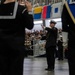 A military ceremony is taking place in a large indoor venue, with a naval officer in dress uniform conducting what appears to be a band or drill team. The central figure wears a white-topped officer's cap and dark dress uniform with gold buttons, arms raised in a conducting gesture. A large audience fills the bleachers in the background, and nautical signal flags are displayed along the upper balcony.