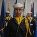 A group of U.S. Navy sailors in dress uniform are standing in formation during what appears to be a formal military ceremony or graduation. The foreground sailor, wearing glasses and a gold aiguillette on the left shoulder, appears to be in a leadership role. Multiple state and military flags are displayed in the background.