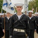 A formation of U.S. Navy recruits in dress uniform stands at attention during what appears to be a graduation or pass-in-review ceremony. The lead recruit in the foreground carries a guidon flag and wears a distinctive white belt with a polished buckle, indicating a leadership or color guard role. The scene takes place outdoors on a military installation with trees and brick buildings visible in the background.