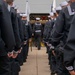 A formation of U.S. Navy sailors in dress blue uniforms and white Dixie cup covers stand at parade rest in two parallel lines, creating a corridor. A single sailor walks between the two ranks toward a building entrance where flags and an audience are visible. The scene appears to be a formal military ceremony or graduation event.