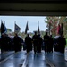 A formation of military personnel, appearing to be U.S. Navy sailors in dress uniforms, marches through a large garage-style bay door into an indoor facility. The procession includes a color guard carrying multiple flags and guidons at the front. The scene appears to be a formal military ceremony or graduation event, photographed from inside looking outward.