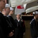 A group of U.S. Navy sailors in dress blue uniforms and white Dixie cup covers are assembled, likely during a formal ceremony or parade formation. A central female sailor with multiple service ribbons and a red lanyard appears to be in a supervisory or senior enlisted role, observing the formation. Flags or pennants are visible overhead, suggesting a ceremonial or official military event.