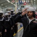 A U.S. Navy military ceremony is taking place inside a large indoor facility, likely a hangar or drill hall. Sailors in dress blue uniforms and white Dixie cup covers are saluting, with the foreground sailor displaying ribbons, a rating badge, and red aiguillette cords indicating a ceremonial role. State and territorial flags are visible hanging from the ceiling in the background.