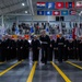 A formal military graduation or pass-in-review ceremony taking place inside a large indoor drill hall or gymnasium. Navy recruits in dress blue uniforms with white sailor caps are marching in formation, led by a guidon bearer. Spectators and family members observe from bleachers in the background, with numerous U.S. state and territorial flags displayed overhead.