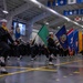 A U.S. Navy recruit graduation ceremony (Pass-in-Review) is taking place inside a large indoor drill hall. Recruits in dress blue uniforms are marching in formation while a color guard carries multiple U.S. state flags. The venue is decorated with state and territorial flags hanging from the ceiling rafters.