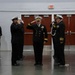 A formal U.S. Navy military ceremony is taking place inside an indoor facility, likely a hangar or gymnasium. Multiple naval officers and enlisted personnel in dress uniforms are rendering salutes simultaneously, suggesting a change of command, retirement, or commissioning ceremony. The central figure appears to be a senior officer receiving honors based on positioning and rank insignia.