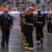 A formal U.S. Navy military ceremony taking place in a large indoor arena or gymnasium. A senior naval officer in dress uniform walks alongside a formation of sailors in dress blues carrying rifles at shoulder arms. A large audience of spectators and seated sailors observes the proceedings from the stands and floor.