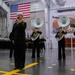 A U.S. Navy military band performs during an indoor ceremony in what appears to be a naval hangar or large facility. Sailors in dress blue uniforms are performing, with one vocalist singing into a microphone while others play sousaphones and a trombone. A large American flag is prominently displayed on the wall in the background, with rows of uniformed sailors standing in formation.