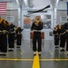 A group of U.S. Navy recruits in dress uniforms are performing a rifle drill routine inside a large indoor facility, likely at a Navy boot camp graduation ceremony. The central figure is tracking a rifle tossed in the air while surrounding recruits handle their own rifles in synchronized positions. The setting features an American flag, a United States Navy flag, and a large naval battle mural on the back wall.