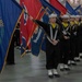 A military color guard ceremony featuring sailors in dress uniforms carrying multiple flags in formation. The personnel are wearing traditional U.S. Navy enlisted dress blue uniforms with white sailor caps and yellow neckerchiefs. The event appears to be taking place inside a large indoor venue such as a hangar or ceremonial hall.