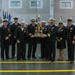 A formal U.S. Navy ceremony taking place in a large indoor facility, likely a drill hall or hangar. A group of naval personnel in dress uniforms are jointly holding a trophy labeled 'Hall of Fame' for a presentation or award ceremony. Rows of uniformed sailors and color guard members with flags are visible in the background.
