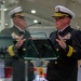 A U.S. Navy flag officer in dress blues stands at a glass podium delivering a speech at what appears to be a formal military ceremony. The officer's reflection is visible in the glass podium surface, creating a mirror-image effect. Multiple military flags and pennants are visible in the background, suggesting an official change of command or commissioning ceremony.