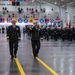 A formal U.S. Navy military ceremony taking place inside a large hangar or drill hall. Two senior naval officers in dress blue uniforms are walking along a marked yellow line, with one officer rendering a salute. Rows of sailors in dress uniforms are standing at attention in the background, flanked by nautical signal flags and observed by spectators in bleachers.