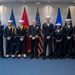 A formal military group photograph featuring one senior naval officer in dress blues with medals and six enlisted sailors in dress uniforms, along with one sailor in a ceremonial/honor guard uniform with gold accoutrements. The group is posed in front of a display of military service flags including the U.S. flag, Marine Corps flag, Navy flag, Air Force flag, and others. The setting appears to be an official Navy facility or ceremonial room.
