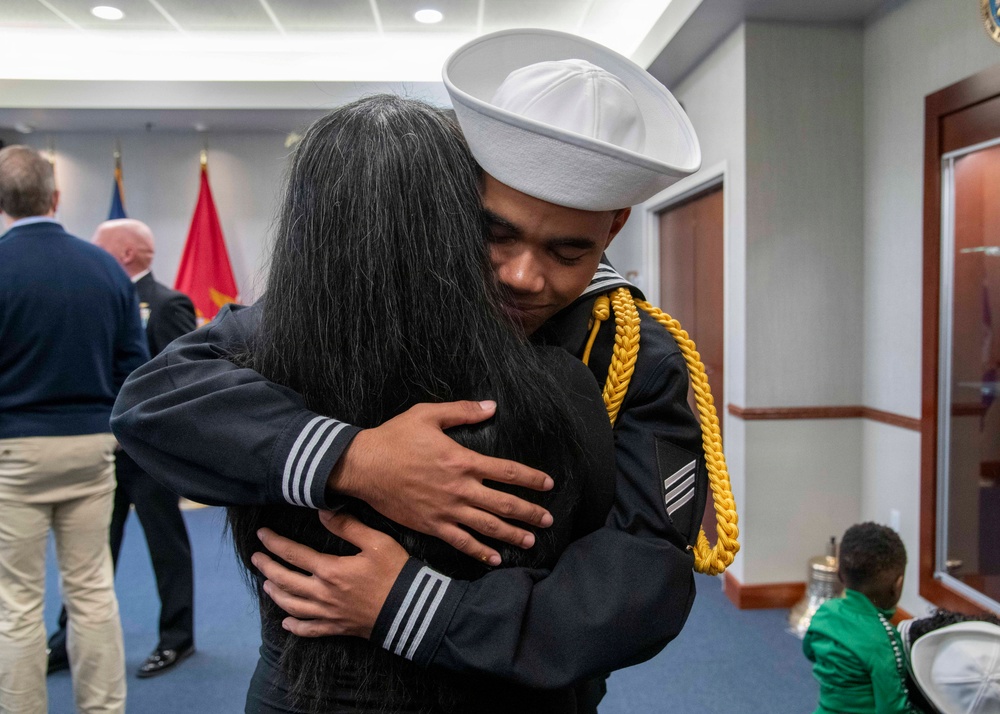 A U.S. Navy sailor in dress uniform, wearing a white Dixie cup cover and displaying gold aiguillette and petty officer rating badges, shares an emotional embrace with a person whose face is not visible. The setting appears to be an indoor military ceremony or event, with flags and other attendees visible in the background. The moment conveys a heartfelt reunion or post-ceremony celebration.