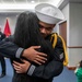 A U.S. Navy sailor in dress uniform, wearing a white Dixie cup cover and displaying gold aiguillette and petty officer rating badges, shares an emotional embrace with a person whose face is not visible. The setting appears to be an indoor military ceremony or event, with flags and other attendees visible in the background. The moment conveys a heartfelt reunion or post-ceremony celebration.