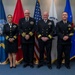 Four U.S. military personnel in Navy dress uniforms pose for a formal group photograph in front of a display of military service flags. The individuals appear to be officers of varying ranks based on their sleeve stripes and insignia. The setting suggests an official military ceremony or formal occasion.