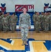 A large group of U.S. Army soldiers in OCP (Operational Camouflage Pattern) uniforms are assembled in formation inside a gymnasium. A senior officer or NCO stands at the front-center of the formation, facing the camera. The backdrop features military unit insignia and signage reading 'IRON' and 'ENGINEERS,' indicating this is likely an Engineer unit formation or official photo.