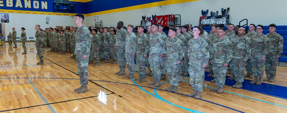 A large formation of U.S. Army soldiers in Operational Camouflage Pattern (OCP) uniforms are standing at attention inside a school gymnasium. The setting appears to be a military ceremony or muster, likely a deployment or mobilization event. The gymnasium belongs to Lebanon school, as indicated by the signage on the walls.