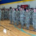 A large formation of U.S. Army soldiers in Operational Camouflage Pattern (OCP) uniforms are standing at attention inside a school gymnasium. The setting appears to be a military ceremony or muster, likely a deployment or mobilization event. The gymnasium belongs to Lebanon school, as indicated by the signage on the walls.