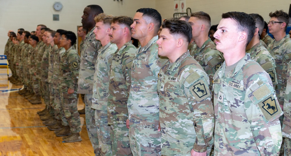 A large formation of U.S. Army soldiers standing at attention in what appears to be a gymnasium or indoor facility. The soldiers are wearing Operational Camouflage Pattern (OCP) uniforms with U.S. Army tape and unit patches visible. The formation appears to be part of a military ceremony or official event.