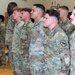 A large formation of U.S. Army soldiers standing at attention in what appears to be a gymnasium or indoor facility. The soldiers are wearing Operational Camouflage Pattern (OCP) uniforms with U.S. Army tape and unit patches visible. The formation appears to be part of a military ceremony or official event.