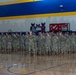 A large formation of military personnel in camouflage uniforms (ACU/OCP pattern) stands at attention inside a school gymnasium. The gymnasium features blue and yellow school colors with 'ENGINEERS' and 'IRON' signage visible on the walls, suggesting this may be a deployment ceremony or military formation event. One soldier appears to be addressing the formation from the front left.