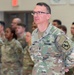 A U.S. Army soldier, identified by the name tape 'TINNER' on their uniform, stands at the forefront of a formation inside what appears to be a military facility. The soldier wears the Operational Camouflage Pattern (OCP) uniform with a unit patch depicting a horse on the right sleeve and a yellow-bordered shoulder sleeve insignia on the left. Multiple soldiers in similar uniforms are visible in the background, standing in formation.