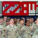 A group of U.S. Army soldiers in OCP (Operational Camouflage Pattern) uniforms stand at attention in a formation inside a military facility. Visible name tapes include Ketner, Sinclair, Keating, Mosley, and Delozier. Behind them is a large red '279th' unit insignia or decorative sign mounted on a wall above bleachers.
