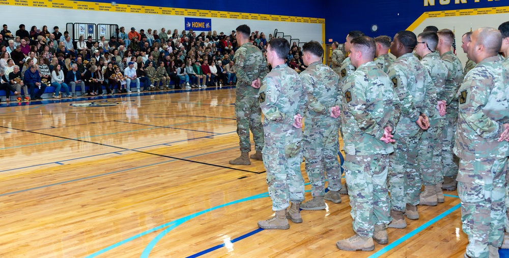 A military ceremony or homecoming event is taking place inside a school gymnasium, with a formation of soldiers in Army Combat Uniforms (ACU/OCP pattern) standing on the basketball court. A large crowd of civilians, including families and community members, fills the bleachers on the left side. The blue and gold gymnasium décor and a 'HOME' banner suggest this is likely a welcome home or send-off ceremony at a school or community facility.