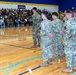 A military ceremony or homecoming event is taking place inside a school gymnasium, with a formation of soldiers in Army Combat Uniforms (ACU/OCP pattern) standing on the basketball court. A large crowd of civilians, including families and community members, fills the bleachers on the left side. The blue and gold gymnasium décor and a 'HOME' banner suggest this is likely a welcome home or send-off ceremony at a school or community facility.