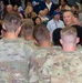A large indoor military formation or ceremony is taking place, with numerous soldiers in US Army OCP (Operational Camouflage Pattern) uniforms standing with their backs to the camera. A civilian man in a light blue shirt appears to be addressing or speaking to the assembled soldiers. A large crowd of civilians, likely family members, is gathered in the background observing the event.